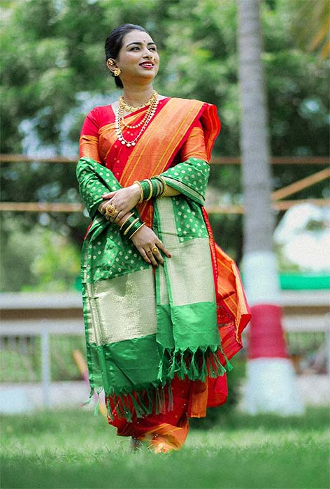 Woman in red Nauvari saree with green pallu and gold jewellery outdoors.